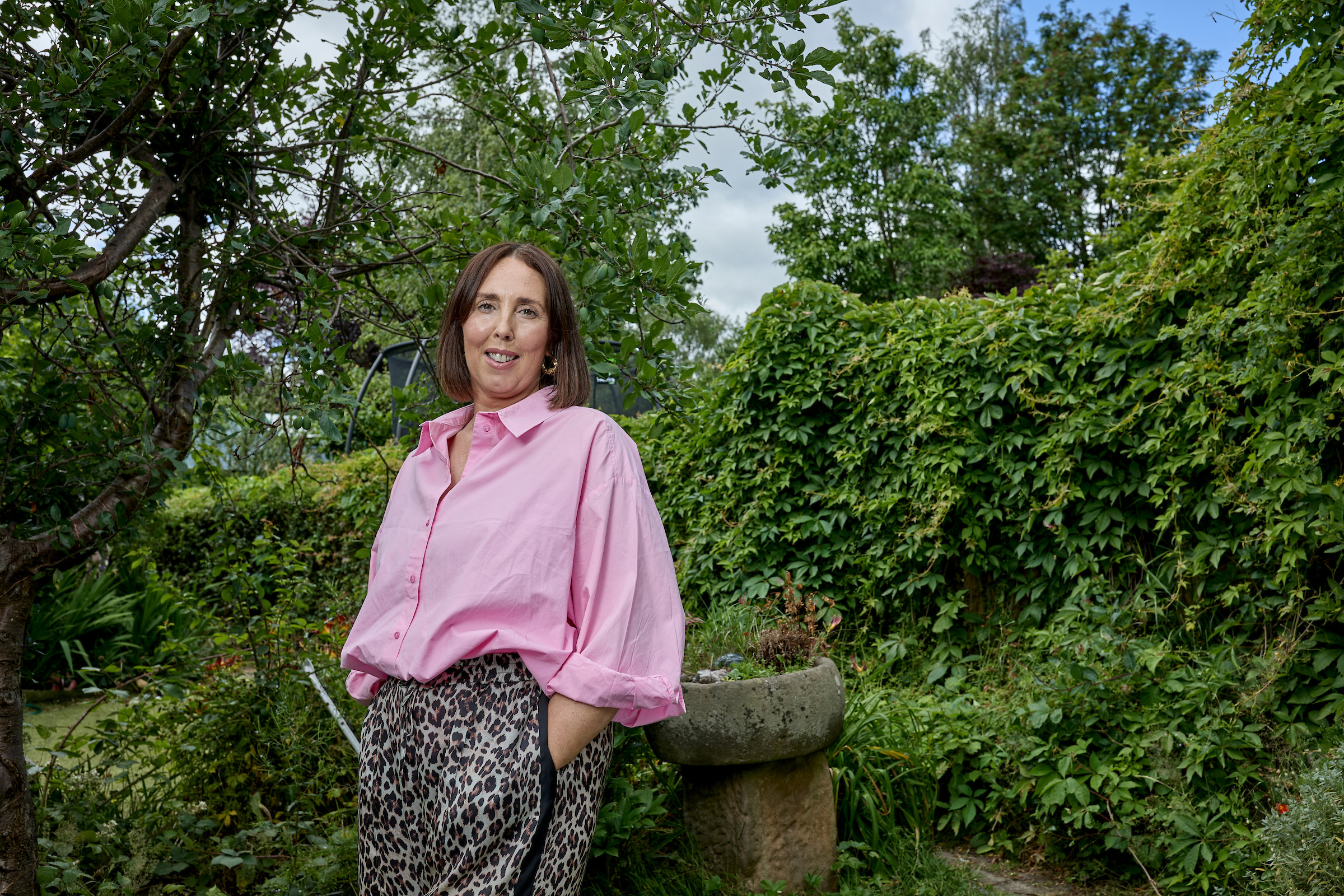 Sally stands in her garden surrounded by greenery. She smiles at the camera and wears a pink top