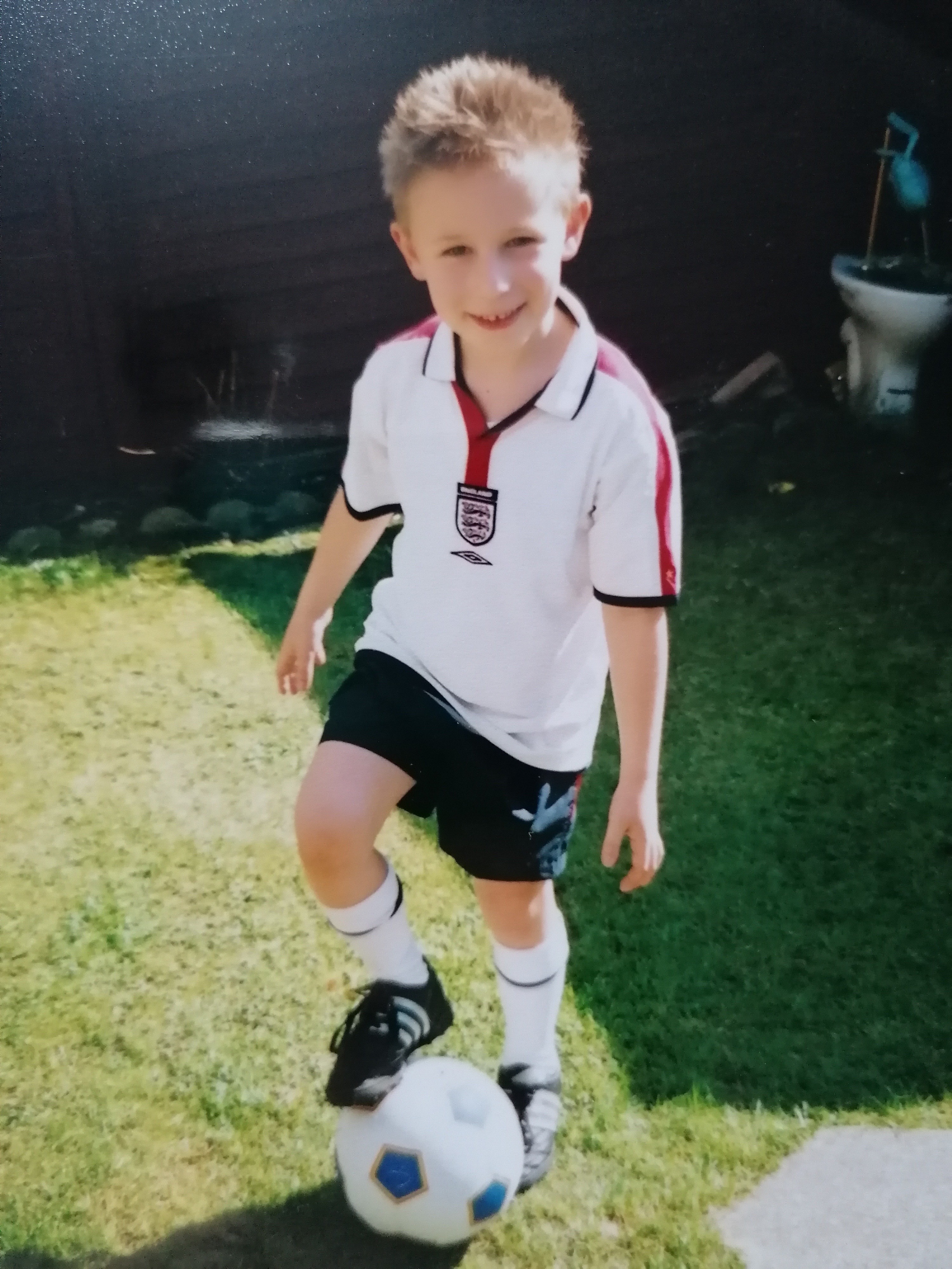 Bradley, a little boy with spiky hair and wearing an England kit, poses with a football