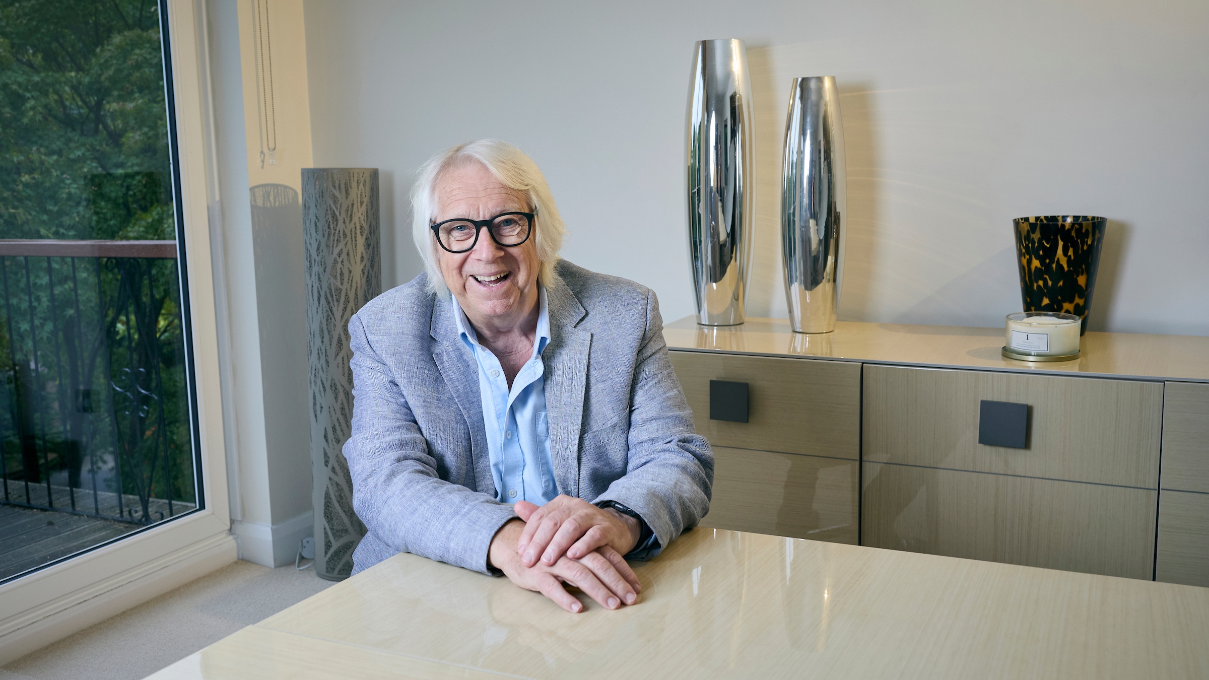 tony sits at his kitchen table in a shirt and blazer, smiling. he has grey hair and black glasses.