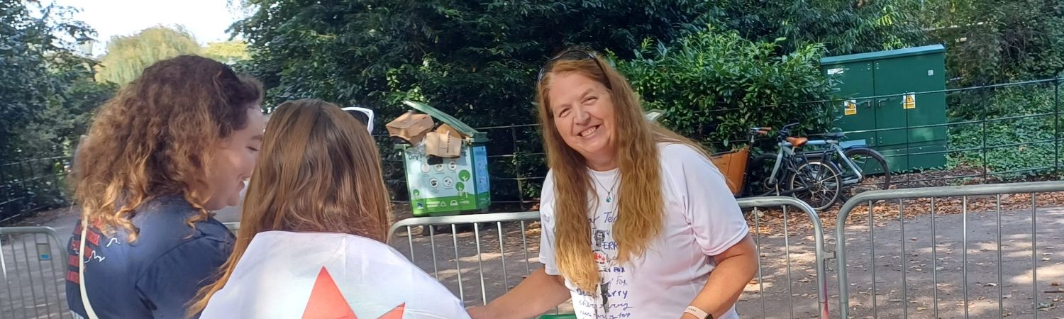 Shelley Battersea 2024 cropped Shelley stands smiling behind a desk outdoors with two people in front - one has a Canadian flag draped around her shoulder