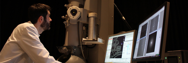 Researcher looking up at electron microscope, surrounded by several screens