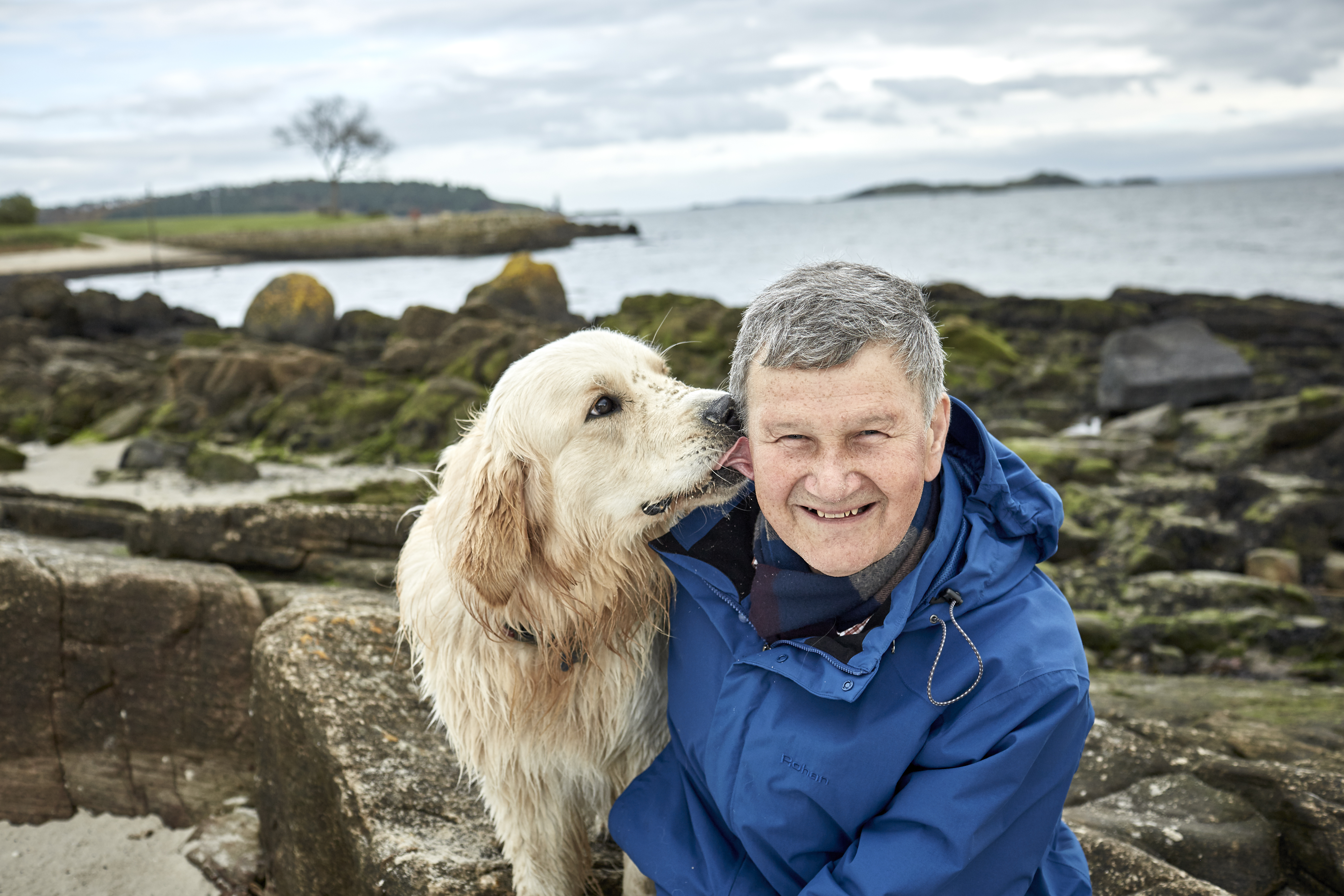 Rob - A grey haired man in a blue jacket sits in front of a lake, smiling at the camera. A golden retriever sits next to him and licks his face