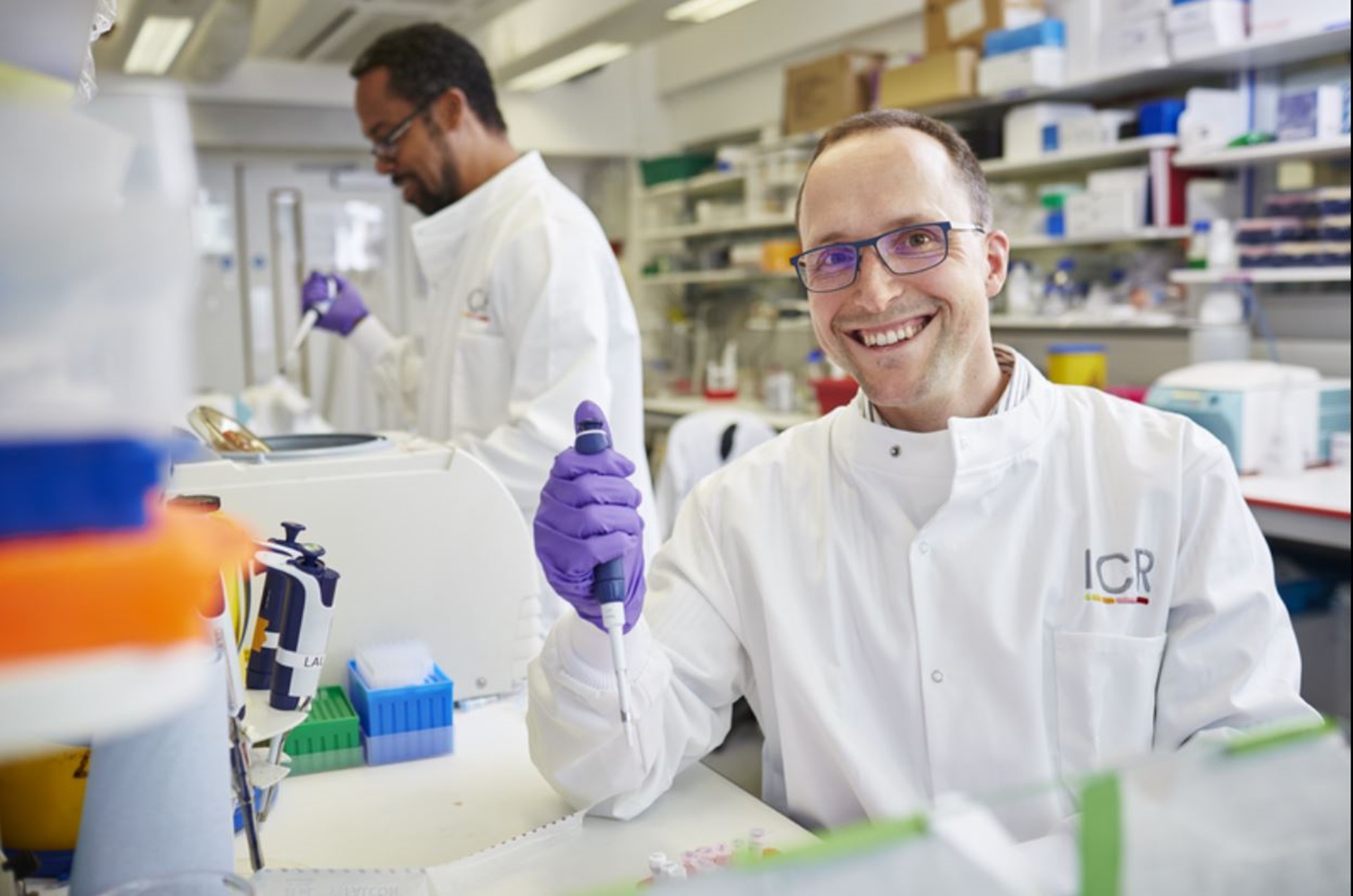 Sebastian - A man wearing purple gloves in a lab coat smiles at the camera. Another scientist in the background is conducting an experiment.